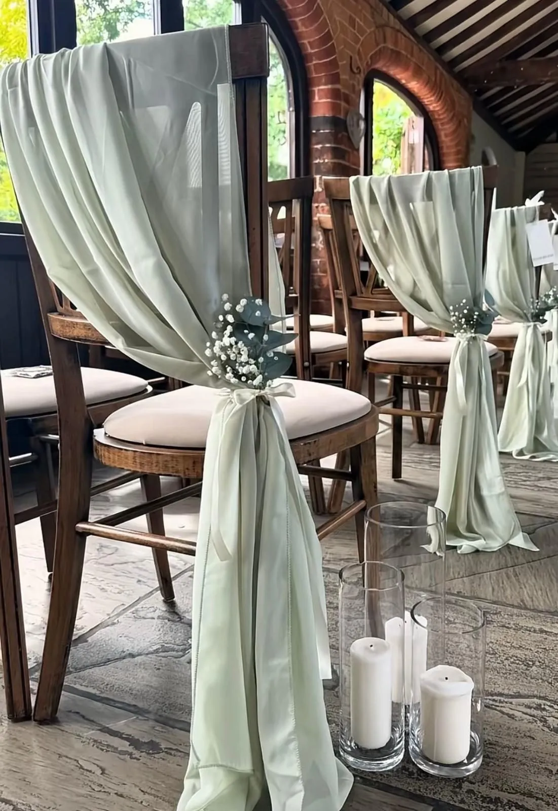 A ceremony aisle styled by NLK: sage green chiffon drapes tied to wooden chairs with gypsophila and ribbon, with pillar candles in glass cylinders along the aisle